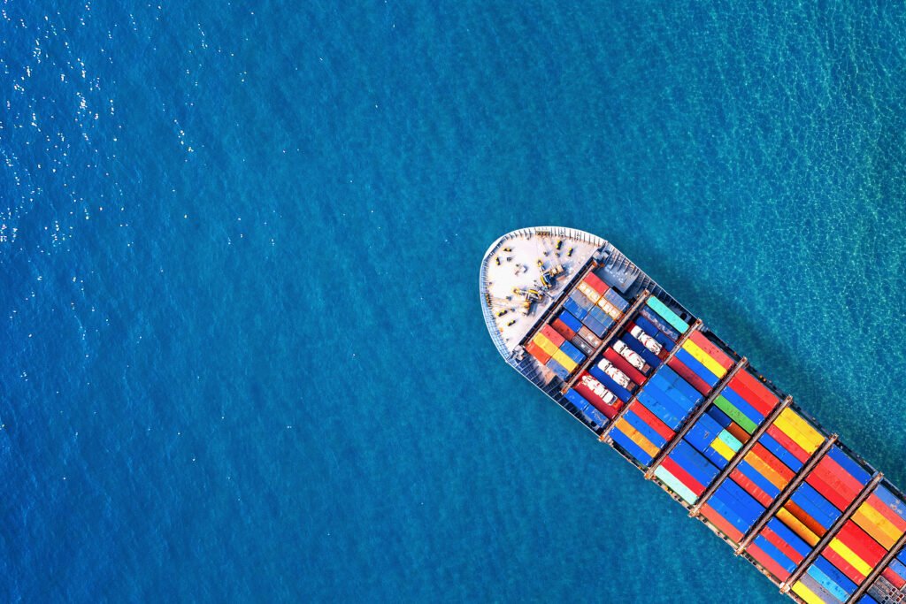 Aerial view of a colorful cargo ship loaded with multicolored shipping containers, sailing through vibrant turquoise-blue water, with the front of the vessel centered in the frame and the vast ocean surrounding it.